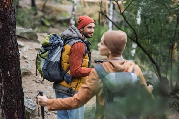 Sonriente hombre con mochila mirando a la mujer trekking en el bosque en primer plano borroso - foto de stock