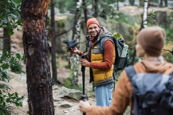 Sonriente hombre sosteniendo la cámara vintage y mirando a la mujer trekking en el bosque en primer plano borroso - foto de stock