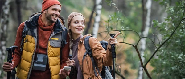 Mujer feliz señalando con el dedo cerca de novio alegre con teléfono inteligente, bandera - foto de stock