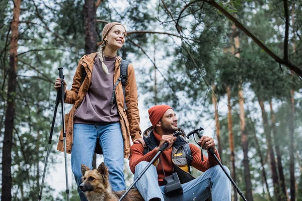 Улыбающаяся пара смотрит в сторону, держа в лесу палки для походов рядом с собакой — стоковое фото