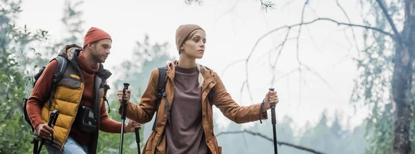 Couple in hats holding hiking sticks while trekking in forest, banner — Stock Photo