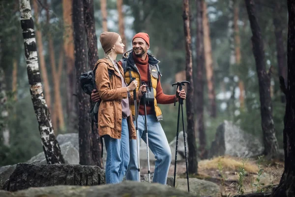 Весела пара в капелюхах тримає пішохідні палички, обіймаючись в лісі — стокове фото