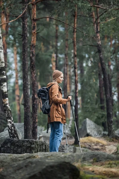 Young woman in hat standing with hiking sticks and backpack in forest — Stock Photo