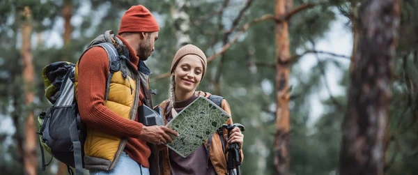 Homem segurando mapa e olhando para mulher alegre com caminhadas varas, banner — Fotografia de Stock