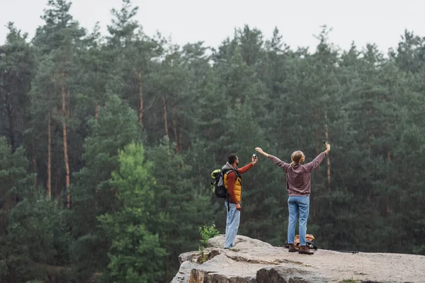 Back view of young couple with smartphones taking selfie on rocky cliff during rain — Stock Photo