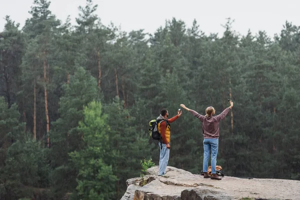 Vista posteriore di giovane coppia con smartphone scattare selfie sulla scogliera rocciosa durante la pioggia — Foto stock