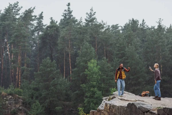 Happy couple with smartphones standing on rocky cliff during rain — Stock Photo