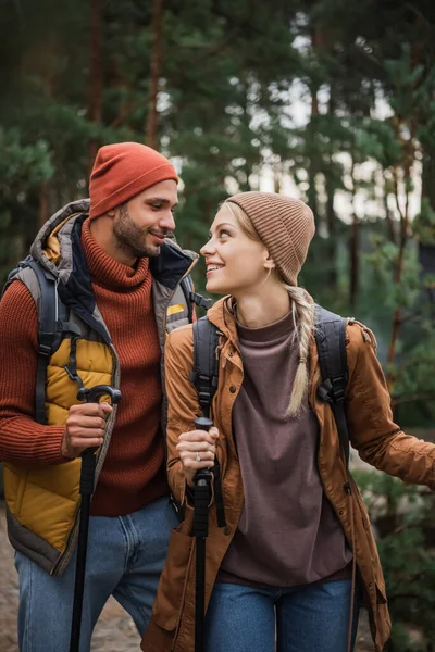 Sonriente joven pareja con mochilas trekking junto con bastones de senderismo y mirándose - foto de stock