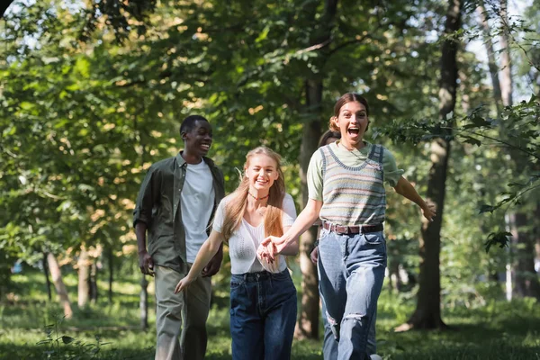 Alegre adolescente niñas corriendo cerca de multiétnicos amigos en parque - foto de stock