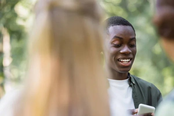 Positivo afroamericano adolescente usando smartphone cerca borrosa amigos - foto de stock