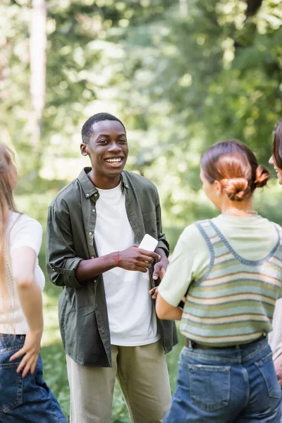 Positive african ameican teenager holding smartphone near friends on blurred foreground — Stock Photo