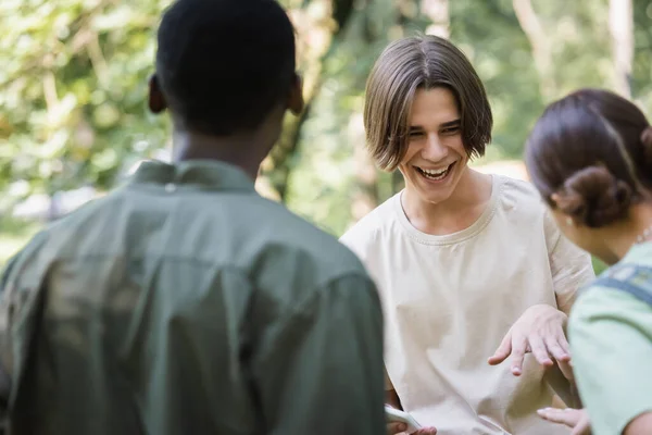 Happy teenager holding cellphone near blurred interracial friends — Stock Photo