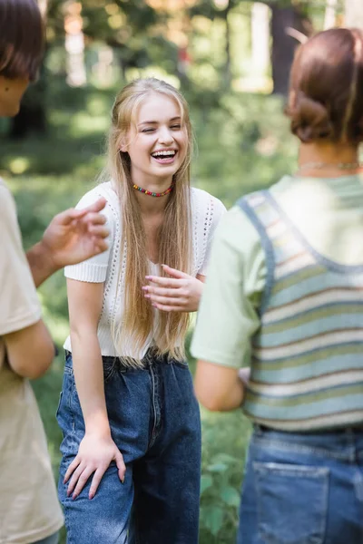 Adolescent souriant avec les yeux fermés debout près des amis sur le premier plan flou dans le parc — Photo de stock