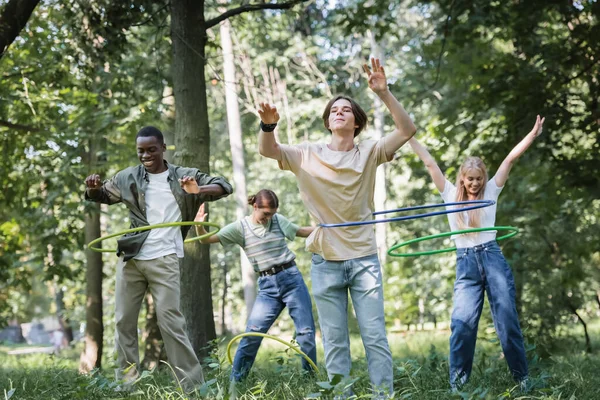 Sonrientes adolescentes multiétnicos retorciendo aros hula en el parque - foto de stock