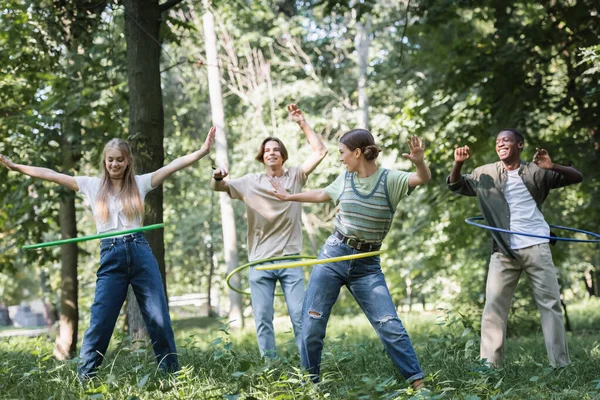 Cheerful multiethnic teenagers twisting hula hoops on grass — Stock Photo