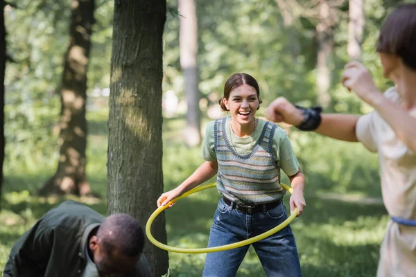 Sonriente adolescente sosteniendo hula hoop cerca de amigos interracial en primer plano borrosa - foto de stock