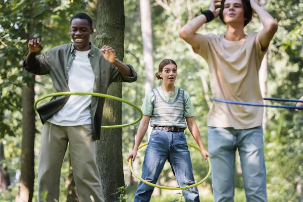 Eccitato ragazza tenendo hula hoop vicino sfocato interrazziale amici — Foto stock