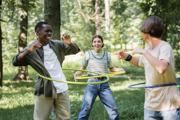 Sonriente chica sosteniendo hula hoop cerca interracial amigos en hierba - foto de stock