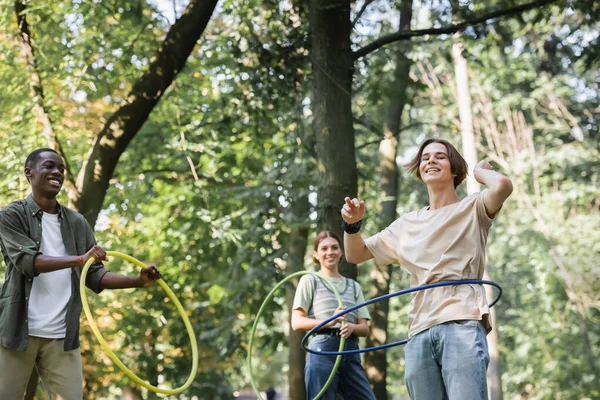 Vista de ángulo bajo de adolescentes interracial torciendo aros hula en el parque - foto de stock