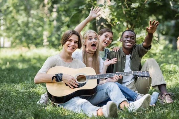 Interracial teenagers with acoustic guitar waving hands in park — Stock Photo