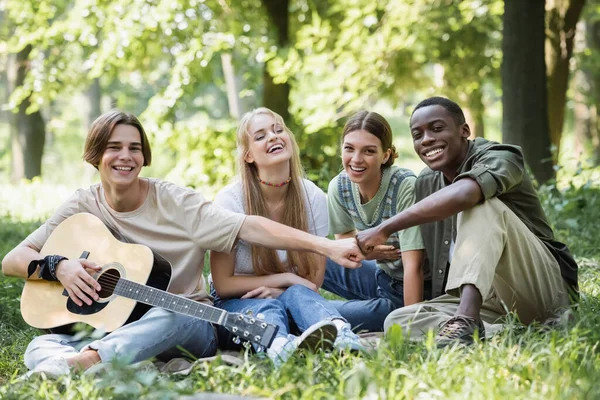 Усміхнені багатоетнічні підлітки з акустичною гітарою роблять кулаки стрибають в парку — стокове фото