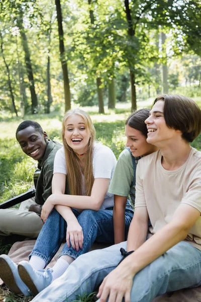 Adolescente alegre amigos sentados perto de menino afro-americano com guitarra acústica no parque — Fotografia de Stock