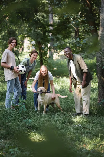 Happy interracial friends with football ball and retriever looking at camera in park — Stock Photo