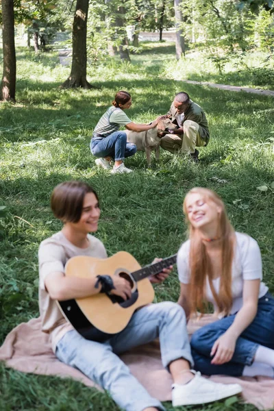 Interracial adolescents caressant récupérateur près de flous amis jouer de la guitare acoustique sur la pelouse — Photo de stock