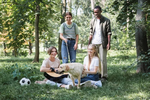 Smiling interracial teen friends spending time with retriever and acoustic guitar — Stock Photo