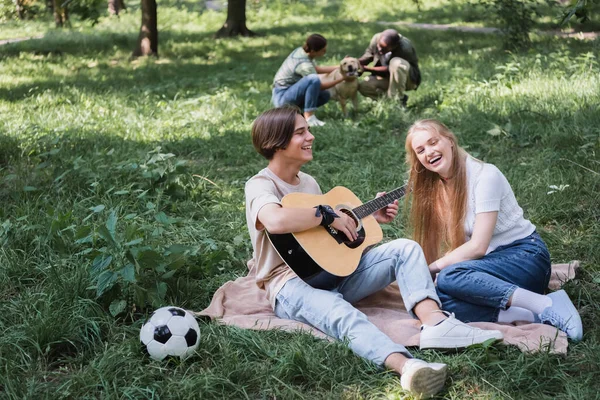 Teenager playing acoustic guitar near friend and football ball on grass — Stock Photo