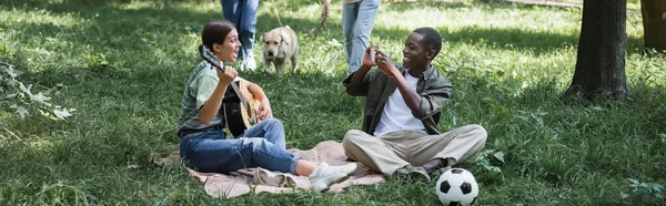 Chica tocando la guitarra acústica cerca de amigo afroamericano con teléfono inteligente en el parque, pancarta - foto de stock