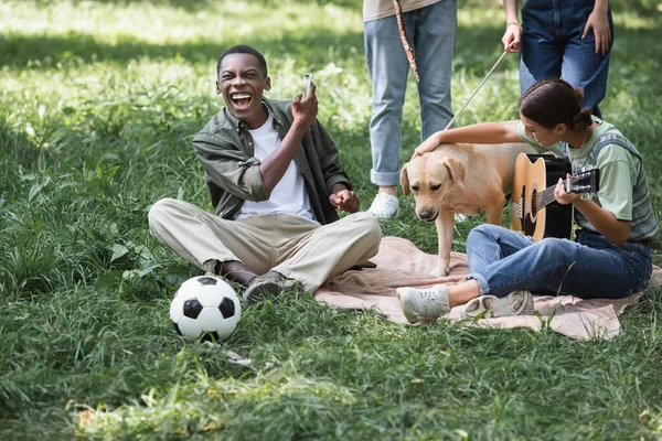 Alegre chico afroamericano sosteniendo teléfono inteligente cerca de amigos con perro recuperador y guitarra acústica en el césped - foto de stock