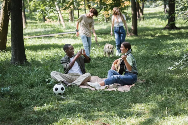 African american teenager using smartphone near friends playing acoustic guitar in park — Stock Photo