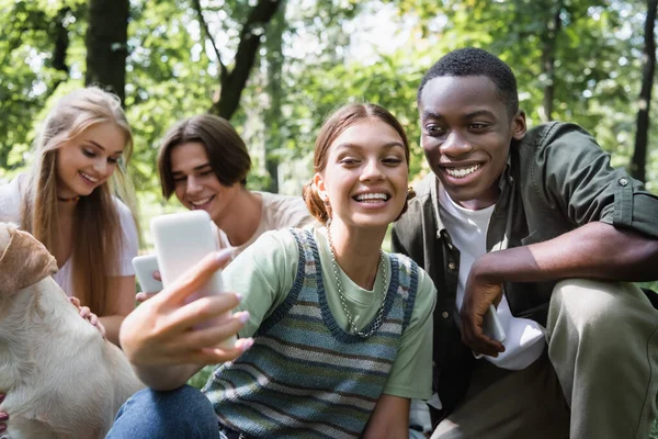 Lächelndes Teenager-Mädchen macht Selfie in der Nähe von afrikanischem amerikanischen Jungen und Freunden mit Retriever im Park — Stockfoto