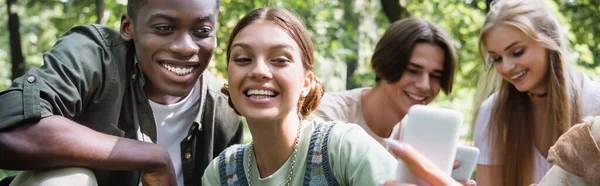 Sonrientes adolescentes interracial tomando selfie cerca borrosa amigos en parque, pancarta — Stock Photo