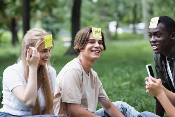 Positive multiethnic teenagers with smartphone playing who i am in park — Stock Photo