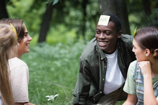 Cheerful interracial teenagers with stickers playing who i am in park — Stock Photo