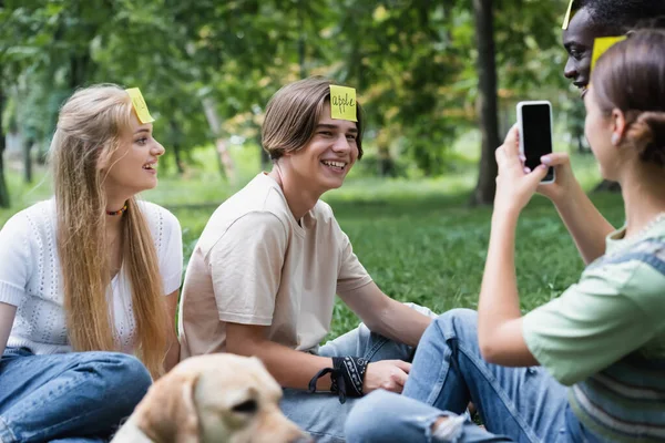Smiling teenagers playing who i am near interracial friends with smartphone and blurred retriever — Stock Photo