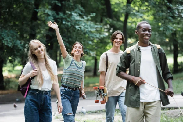 Sorridente adolescente che saluta la fotocamera vicino agli amici interrazziale nel parco — Foto stock