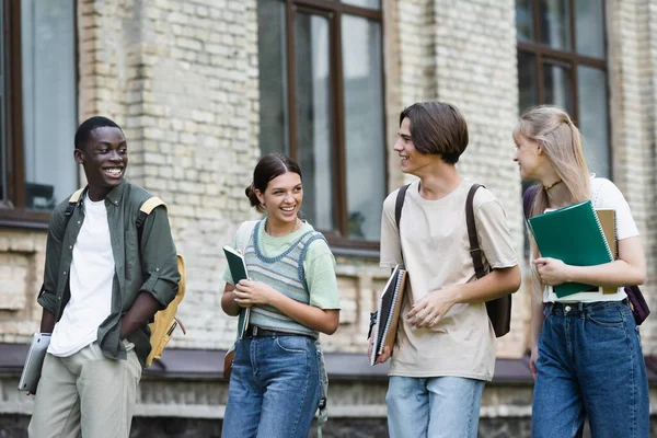 Smiling multiethnic students with notebooks and laptop walking near building — Stock Photo