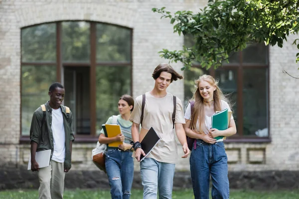 Cheerful students with backpacks and notebooks walking near blurred multiethnic friends — Stock Photo