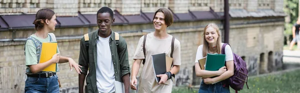 Smiling student talking to interracial friends with backpacks outdoors, banner — Stock Photo