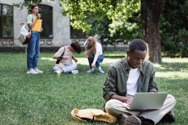 Estudante afro-americano usando laptop perto de mochila e amigos embaçados na grama — Fotografia de Stock