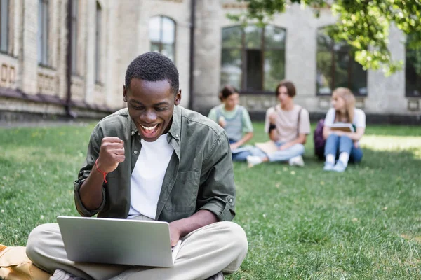 Aufgeregte afrikanisch-amerikanische Studentin zeigt Ja, während sie Laptop auf dem Rasen benutzt — Stockfoto
