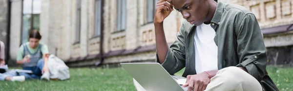Estudiante africano americano pensativo usando el ordenador portátil al aire libre, bandera - foto de stock