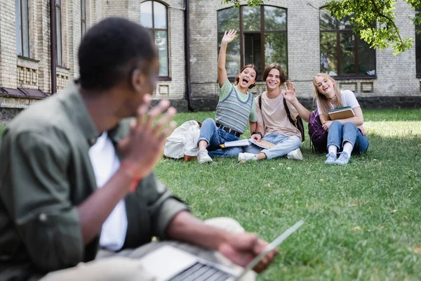 Cheerful students waving hands at blurred african american friend on lawn — Stock Photo