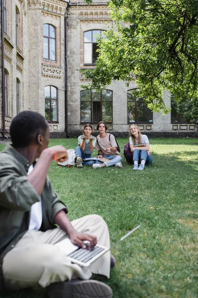 Étudiants souriants regardant un ami afro-américain flou avec ordinateur portable à l'aube — Photo de stock