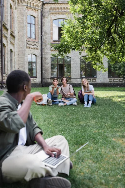 Lächelnde Studenten schauen verschwommenen afrikanisch-amerikanischen Freund mit Laptop an — Stockfoto