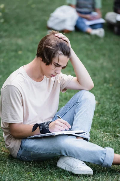 Estudiante escribiendo en el cuaderno mientras está sentado en la hierba - foto de stock