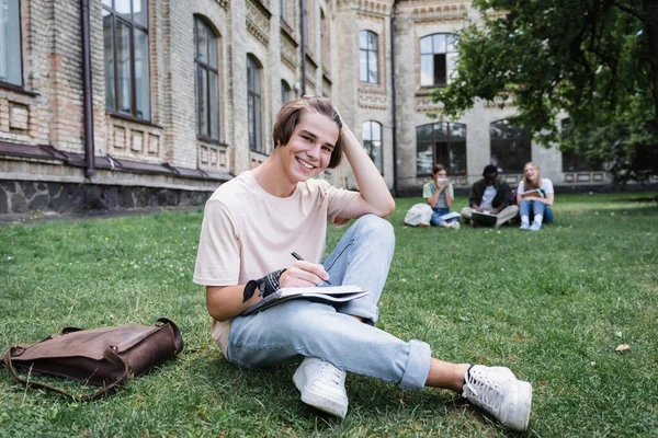 Cheerful student writing on notebook and looking at camera on lawn — Stock Photo